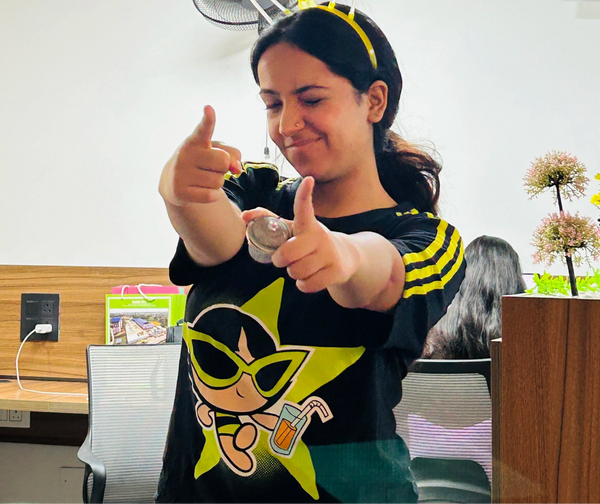 A woman in a black t-shirt with a yellow star graphic and a matching yellow headband with clear plastic straws, posing with two thumbs pointing forward in an office setting.